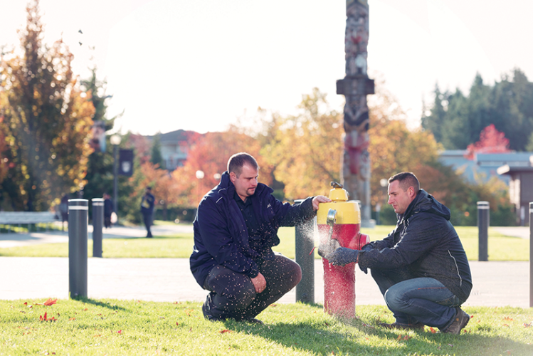 Roger Cerny and Paul McLaughlin testing at a campus hydrant by UBC’s reconciliation pole. Image Credit: Martin Dee / University of British Columbia.